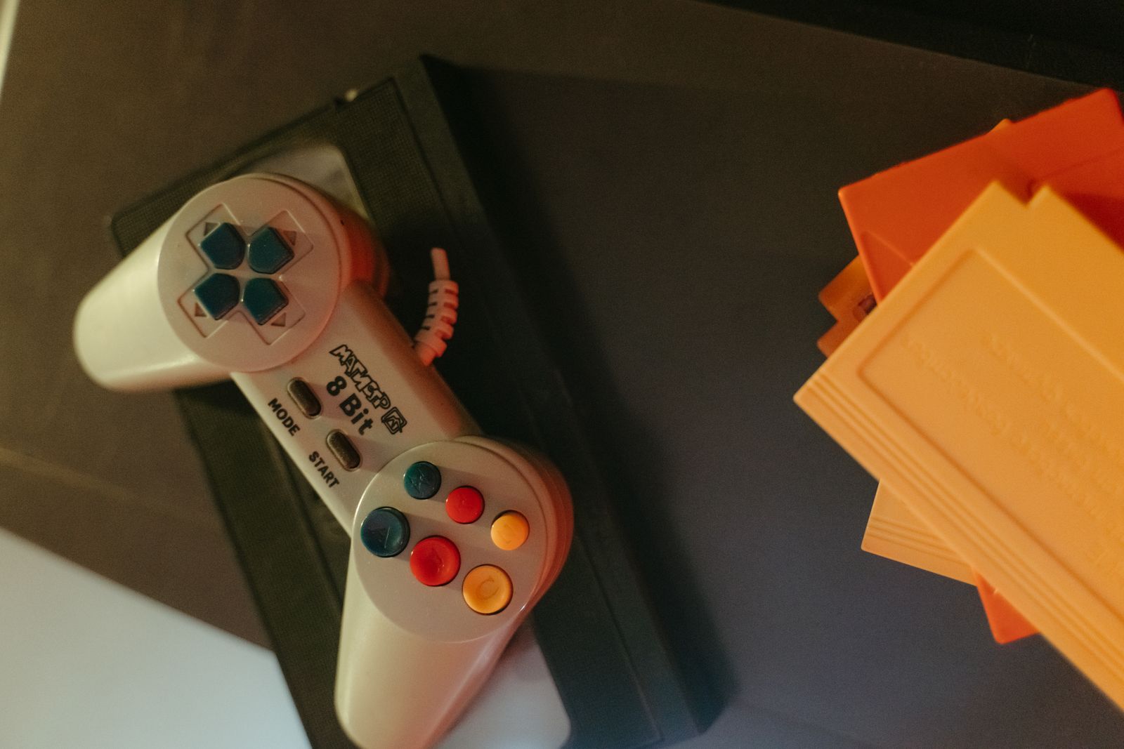 薄暗い机の上に置かれた8ビット時代のゲームパッドと、積み重ねられたオレンジ色のカートリッジ